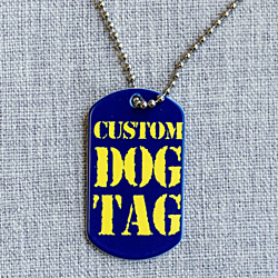 A dark blue military dog tag with yellow text that reads, "custom Dog Tag". The tag is pictured with a ball chain running through a hole in the top of the tag. This dog tag is shown on a gray fabric background. 