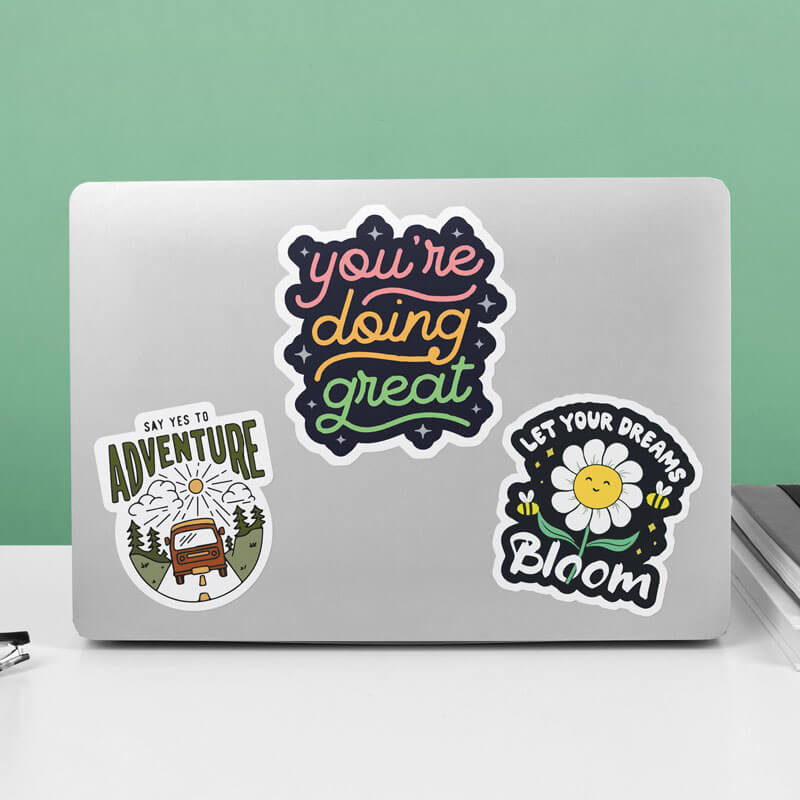 Laptop decorated with three colorful vinyl stickers: "You're doing great" in rainbow lettering, "Say Yes to Adventure" with a camper van and mountains, and "Let Your Dreams Bloom" with a smiling flower and bees.