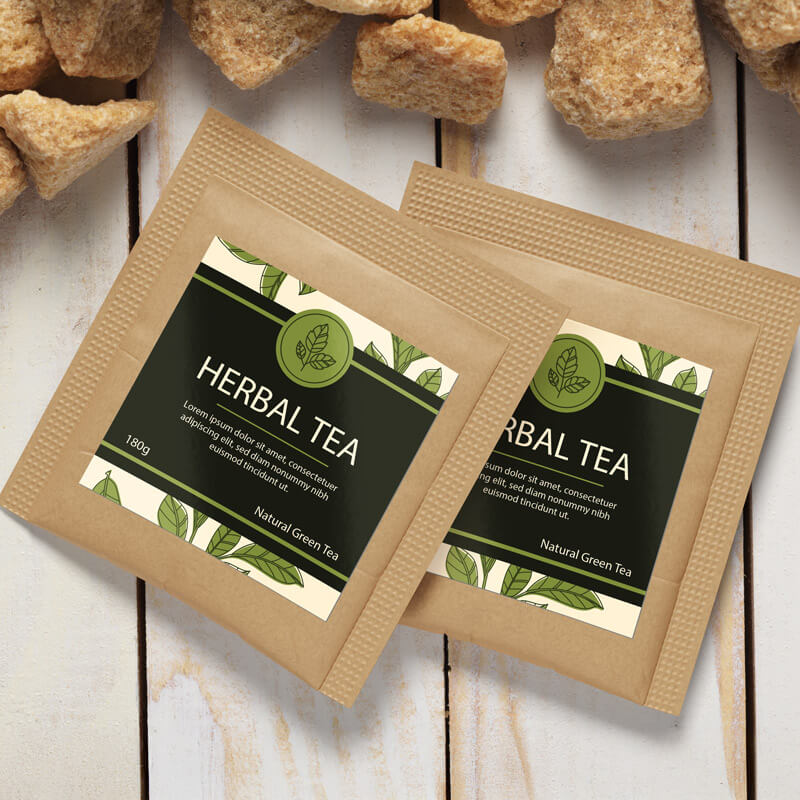 Two square labels with green leaf illustrations and the words "Herbal Tea" are placed on natural kraft paper tea pouches, resting on a wooden surface with raw sugar cubes.