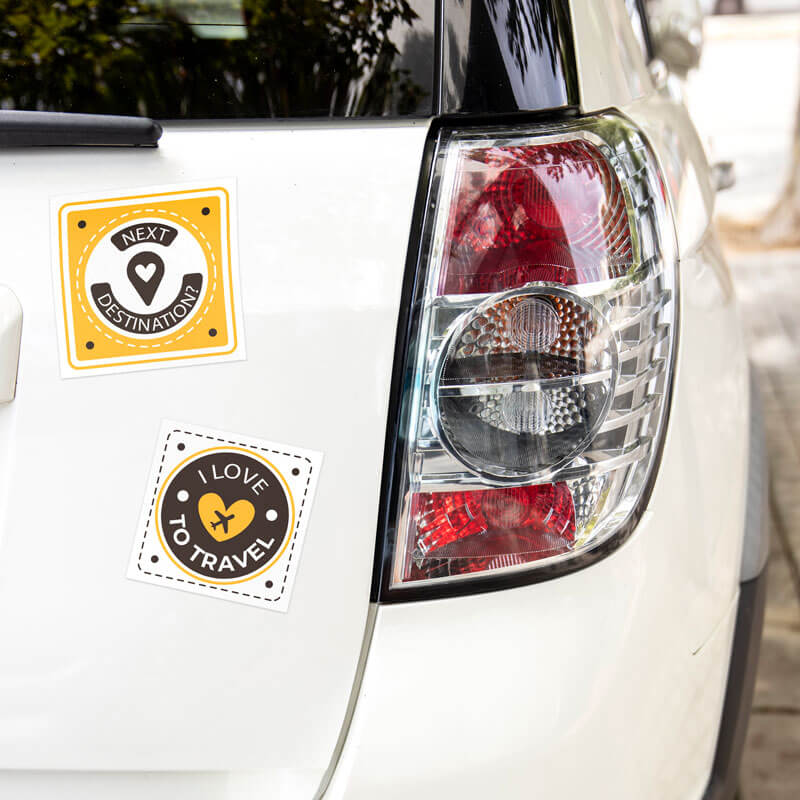 Two travel-themed square stickers, one reading "Next Destination" in yellow and the other "I Love to Travel" in brown and yellow, are placed on the rear of a white car near the taillight.
