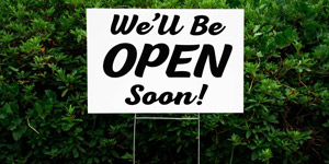 A yard signs promoting a store opening soon installed in front of some bushes.