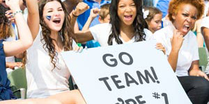 Three woman at a sporting event holding up a corrugated sign cheering on their favorite team.